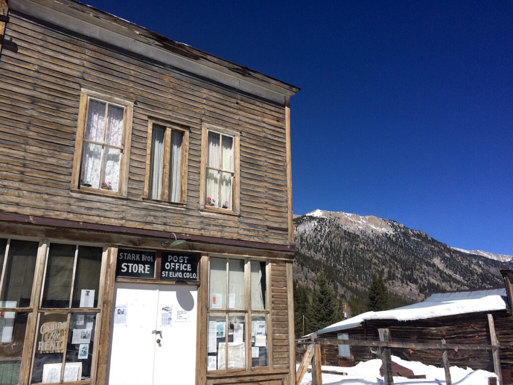 Wood-shingled mining-era building in ghost town with snowy mountains in background on sunny spring day. St. Elmo Ghost Town, near Buena Vista and Salida, Colorado.