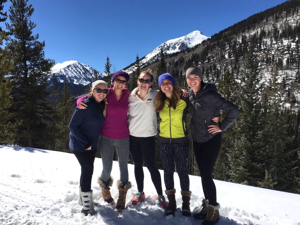 Group of women smiling at camera on snowy trail with snow-capped mountain views and pine trees in background on sunny spring day. Winter hike near St. Elmo Ghost Town, near Buena Vista and Salida, Colorado.