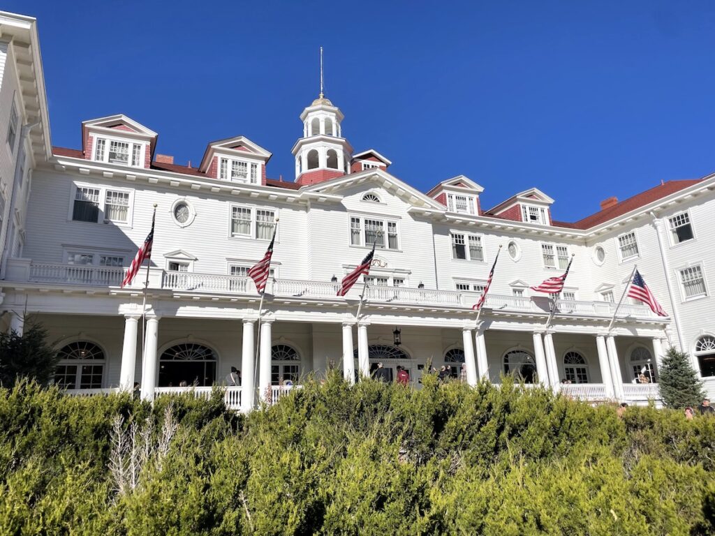 Historic hotel with white siding, red trim, American flags, and green hedges on sunny day. Stanley Hotel, Estes Park, Colorado.