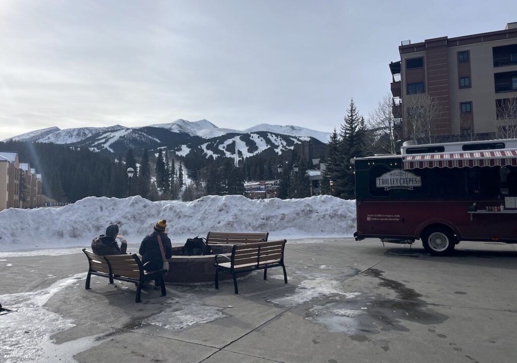 Benches around fire pit next to food truck trolley overlooking mountain views on winter day. Trolley Crepes, Breckenridge Village Resort, Breckenridge, Colorado.