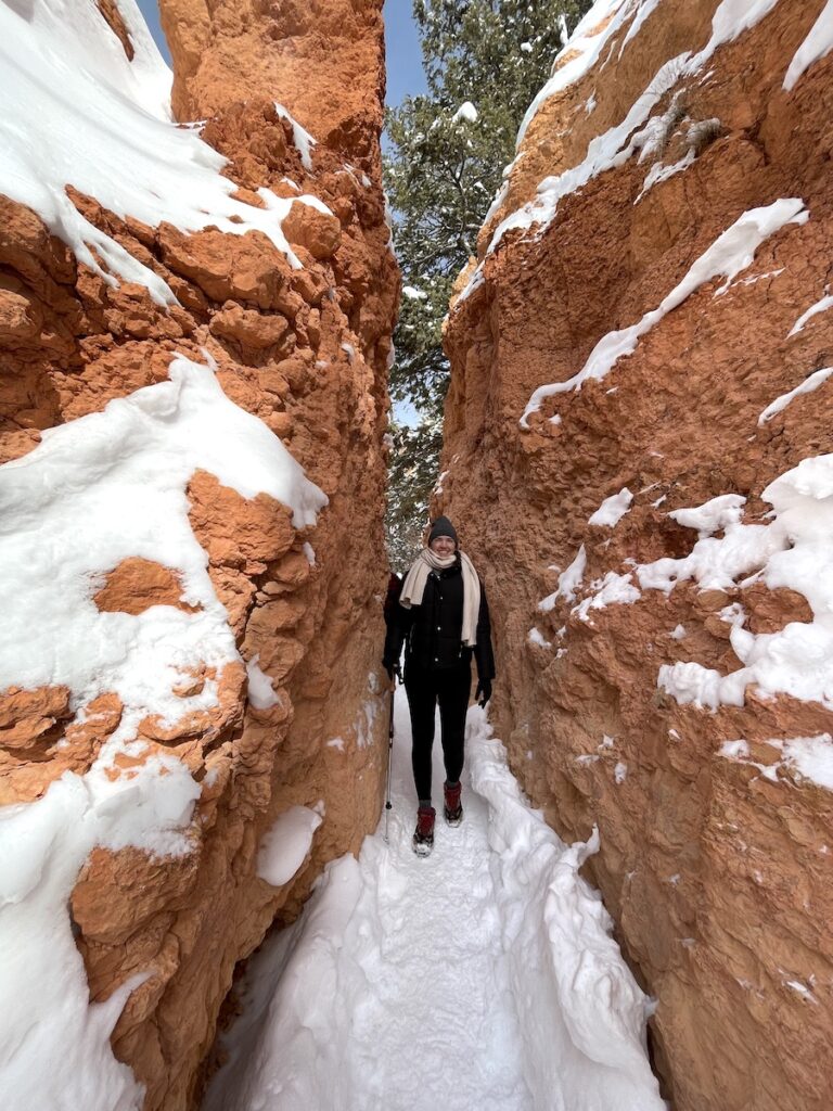 Woman dressed warmly for winter hiking through narrow red rock opening surrounded by snow. Queens Garden via Navajo Loop Trail, Bryce Canyon National Park, Utah in winter.