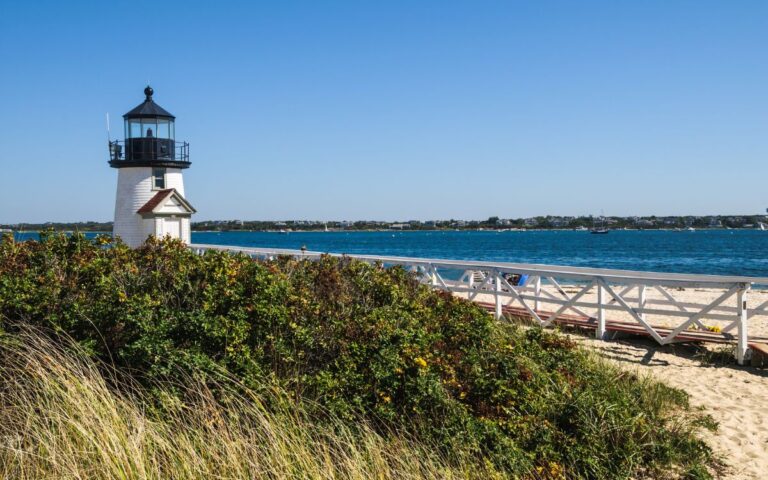 Small white lighthouse with black top on sandy beach with wooden dock leading to lighthouse entrance, lighthouse overlooks Atlantic Ocean with sailboats in the distance. Brant Point Lighthouse, Cape Cod, Nantucket, Massachusetts, New England.