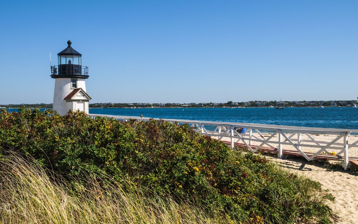Small white lighthouse with black top on sandy beach with wooden dock leading to lighthouse entrance, lighthouse overlooks Atlantic Ocean with sailboats in the distance. Brant Point Lighthouse, Cape Cod, Nantucket, Massachusetts, New England.