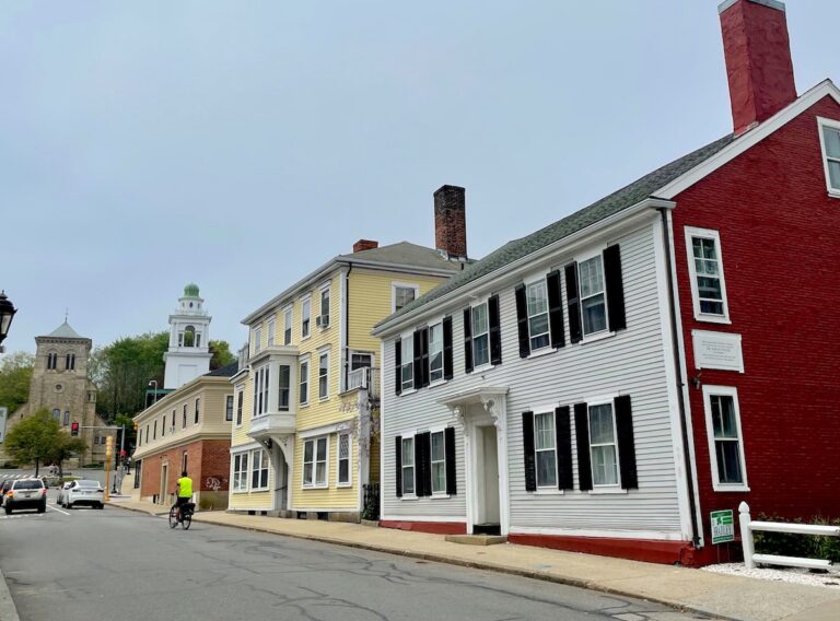 Narrow roadway lined with old, colorful homes with church in distance. Historic homes on Leyden Street, Plymouth, Massachusetts.