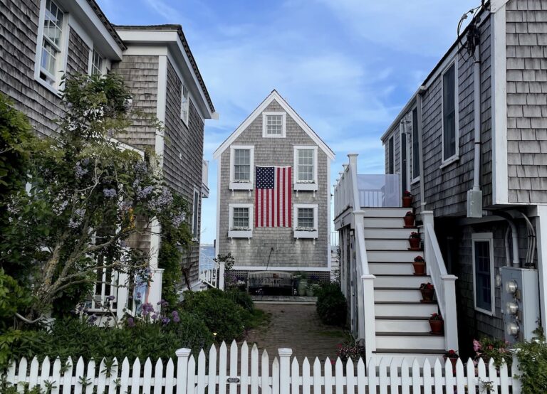 Small shingled, grey house behind white picket fence with huge American flag hanging from side of house. Provincetown, Cape Cod, Massachusetts, New England.