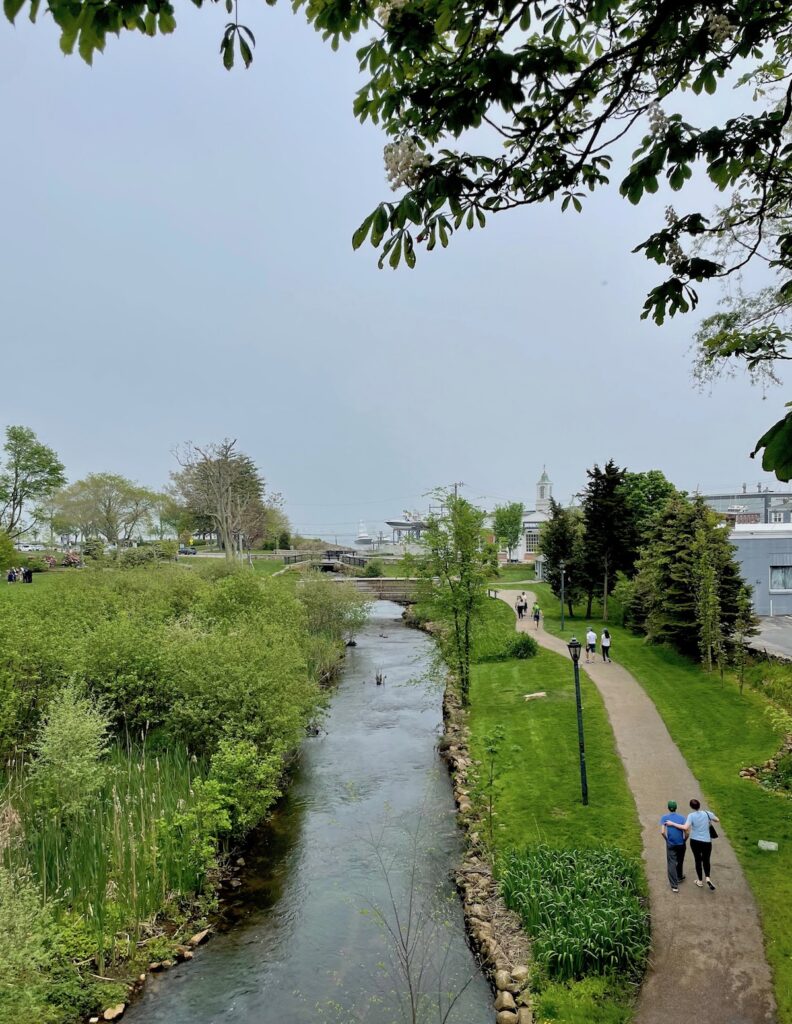 People walking along paved path following narrow stream and gardens on gloomy day. Brewster Gardens, Plymouth, Massachusetts.