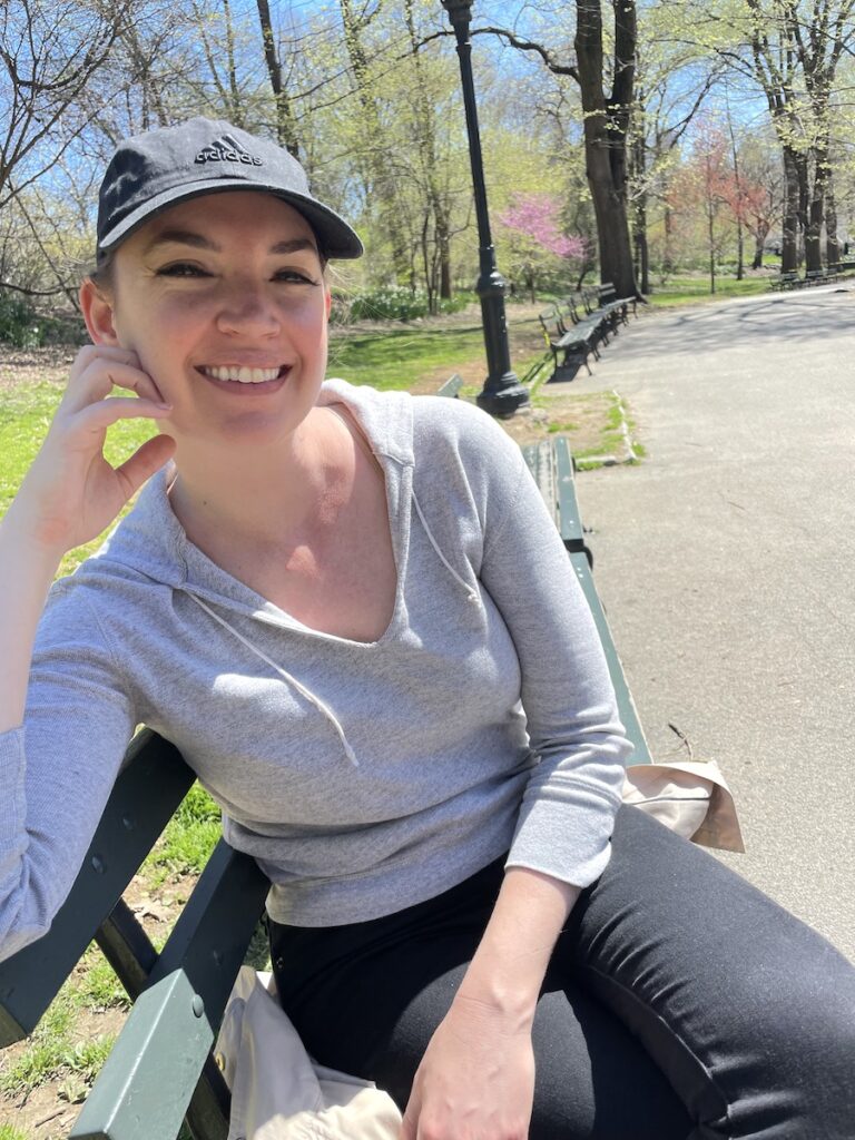 Woman on park bench with budding green and pink trees in background on sunny spring day. Central Park, Manhattan, New York City, New York.