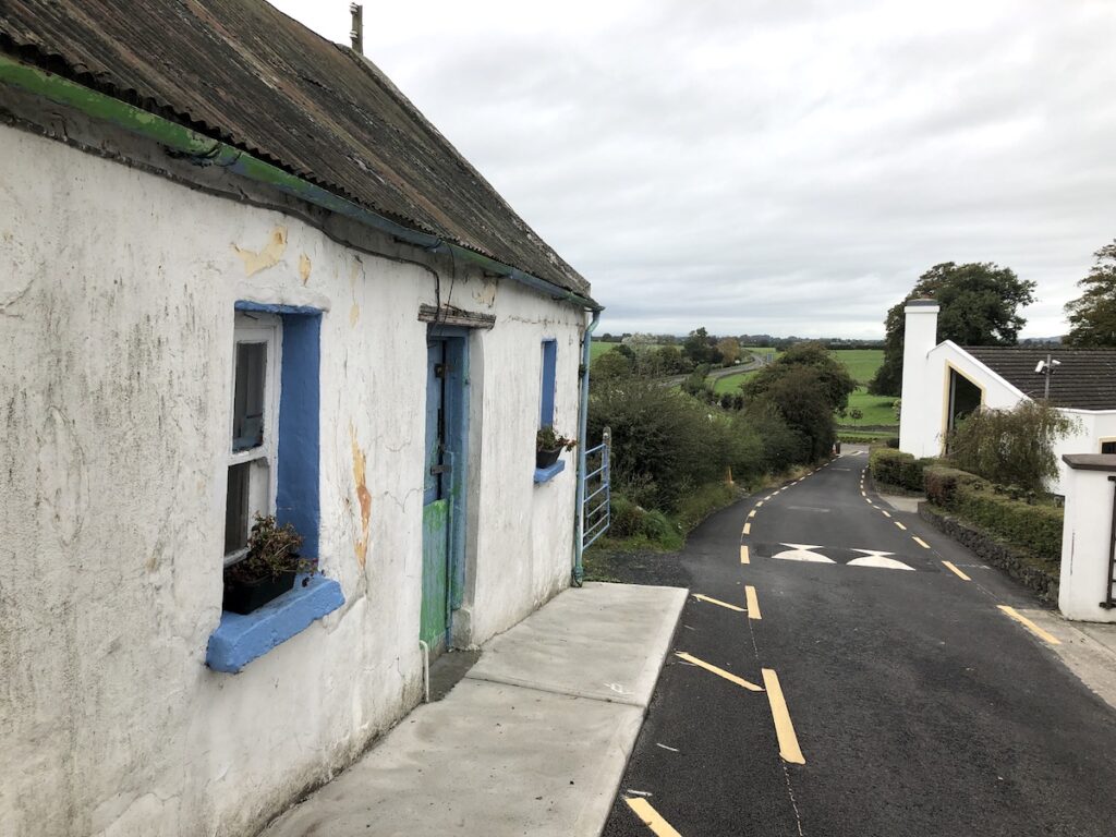 Old, small white house with blue trim sitting right on narrow one-lane road with green hills in background on gloomy day. Irish Countryside, near Rock of Cashel, Ireland road trip.