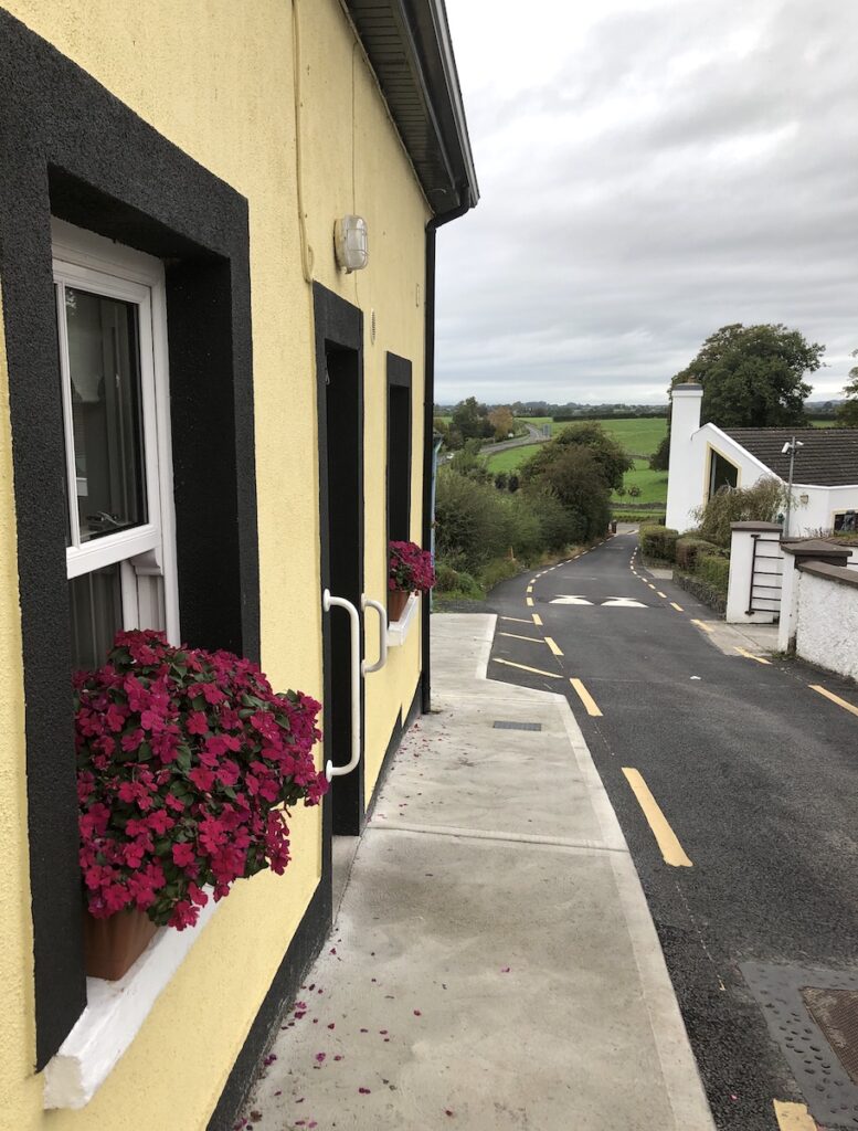 Small yellow house with black trim and pink flowers, sitting right on narrow one-lane road with green hills in background on gloomy day. Irish Countryside, near Rock of Cashel, Ireland road trip.