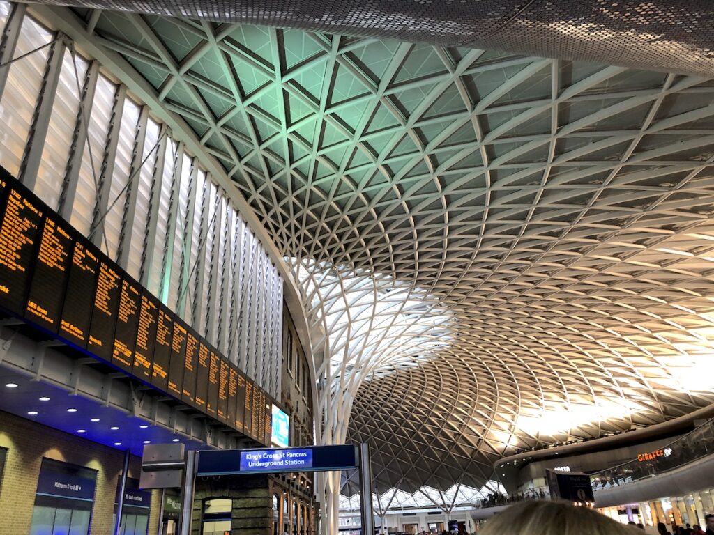 Train station with large geometric ceiling art above large screen with train schedule. King's Cross Railway Station, London, England, UK.