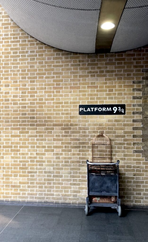 Brick wall with half of a luggage trolley cart attached to wall under sign reading "Platform 9 3/4." Platform 9 3/4, King's Cross Train Station, London, England, UK.