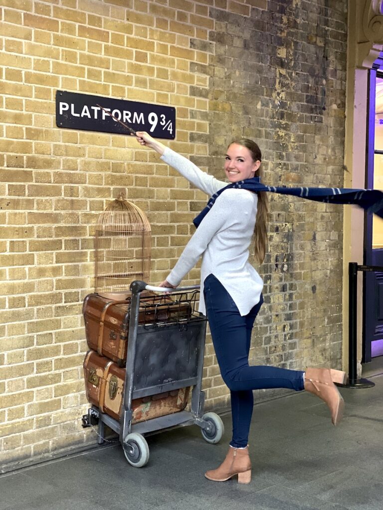 Woman smiling in Ravenclaw scarf holding wand and trolley under sign reading "Platform 9 3/4." Harry Potter's Platform 9 3/4, King's Cross Station, London, England, UK.