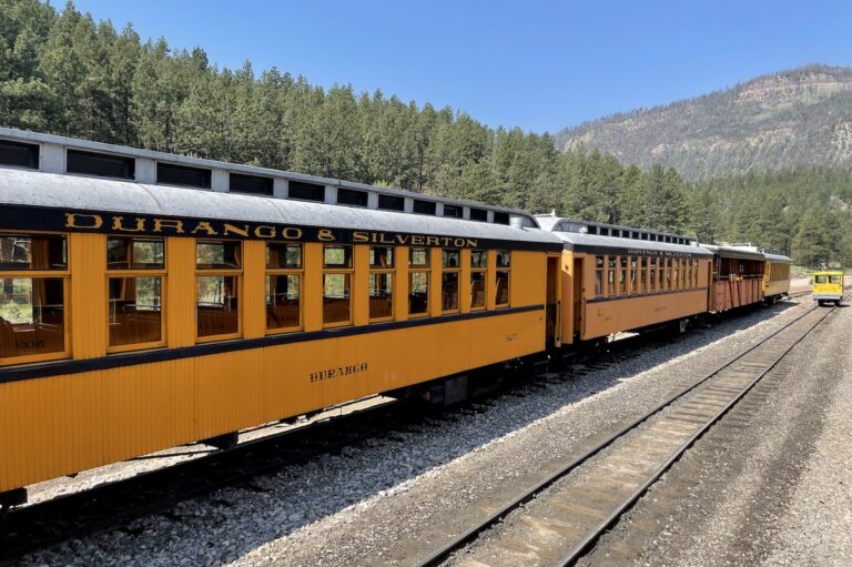 Orange open-air historic train cars in mountains surrounded by pine trees on sunny summer day. Durango & Silverton Narrow Gauge Railroad, San Juan Mountains of the Rocky Mountains, Durango, Colorado.