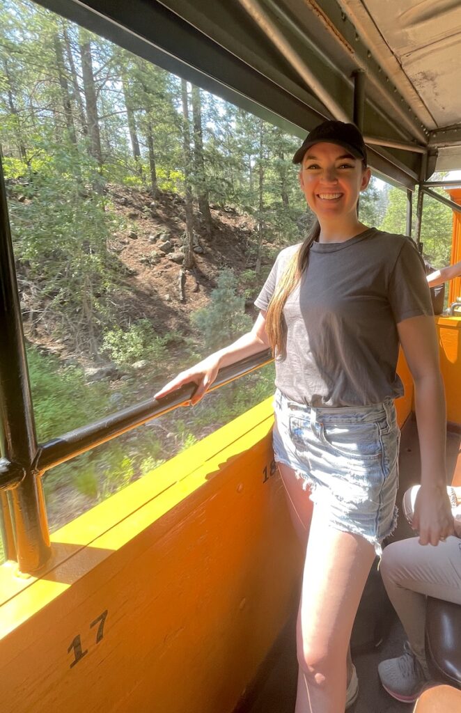 Woman standing next to window of open air train car during train ride through the mountains in summer. Durango & Silverton Narrow Gauge Railroad, Cascade Canyon Route, San Juan Mountains of the Rocky Mountains, Durango, Colorado.