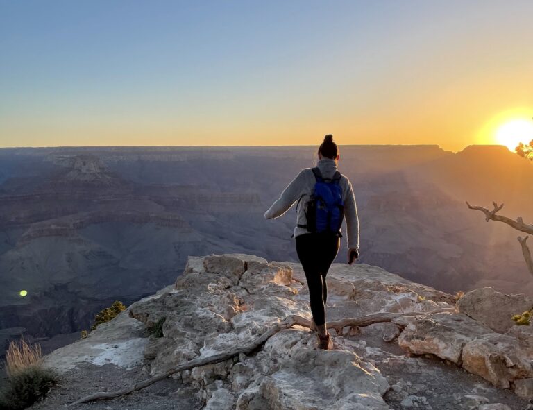 Woman on rocky hiking trail overlooking red, orange, and purple Grand Canyon during sunrise on cloudless, but cold spring morning. Sunrise at Yaki Point, Grand Canyon National Park South Rim, Arizona.