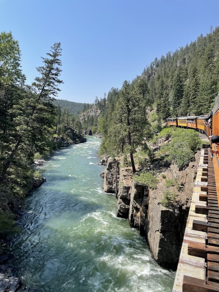 Orange train cars of historic steam train crossing wooden bridge over blue Animas River surrounded by pine trees in mountains on sunny summer day. Durango & Silverton Narrow Gauge Railroad, San Juan Mountains of the Rocky Mountains, Durango, Colorado.