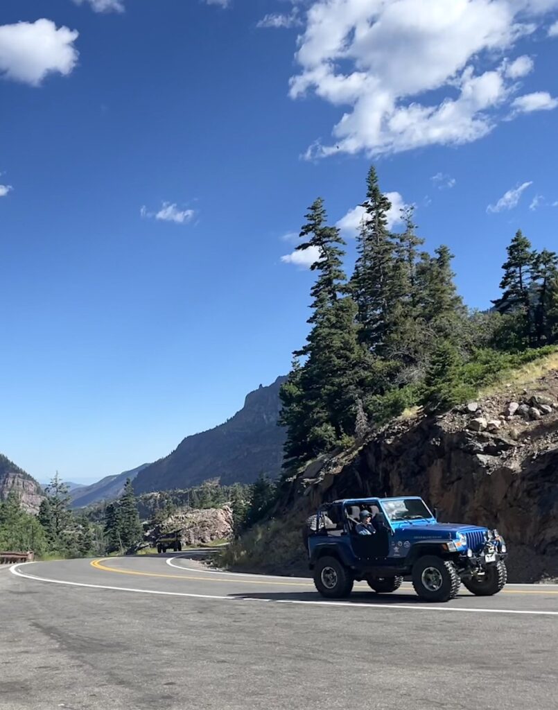 Blue Jeep driving winding mountain road on sunny summer day with pine trees and mountain views in background. Million Dollar Highway US 550 near Ouray and Silverton, Colorado.