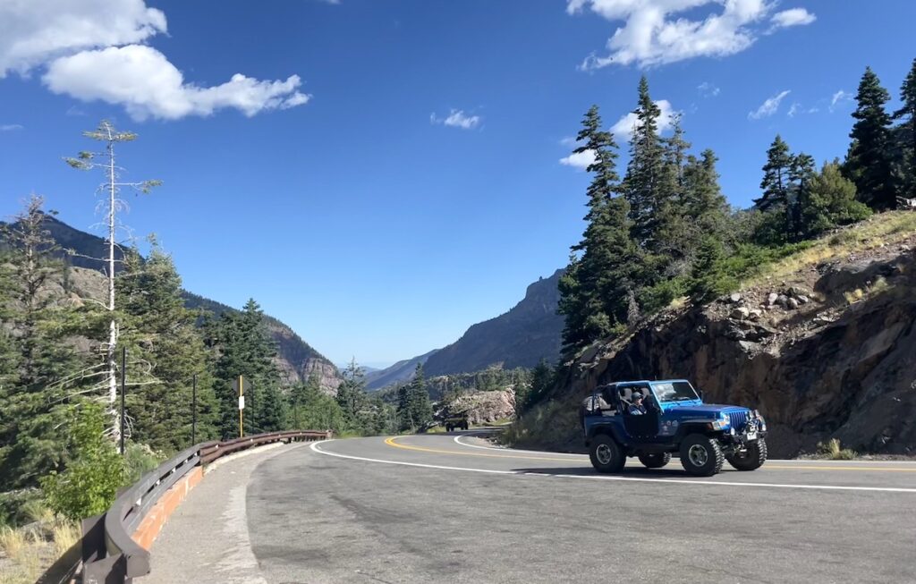 Blue Jeep driving winding mountain road on sunny summer day with pine trees and mountain views in background. Million Dollar Highway US 550 near Ouray and Silverton, Colorado.
