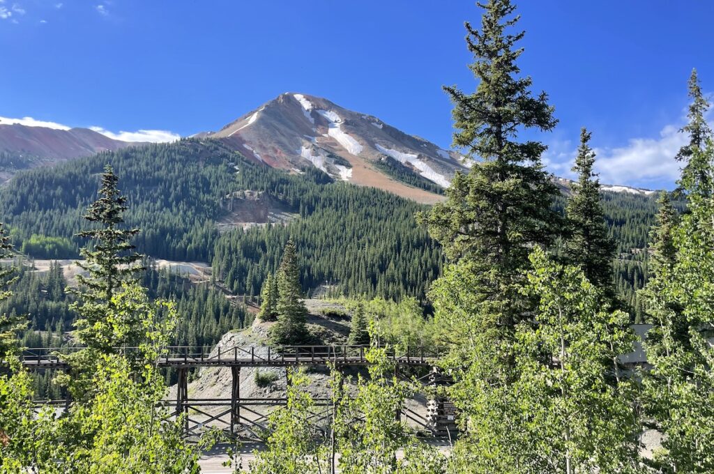 Tall, red mountain with green trees on sunny summer day, in foreground is old mining equipment. Red Mountain Overlook, US 550, Million Dollar Highway, near Silverton and Ouray, Colorado.