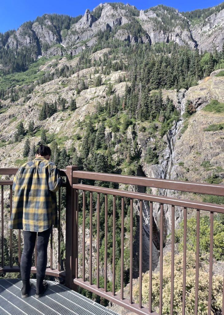 Woman looking out overlook with tall skinny waterfall in mountains in distance on sunny summer day. Bear Creek Falls Overlook, Million Dollar Highway, near Ouray and Silverton, Colorado.
