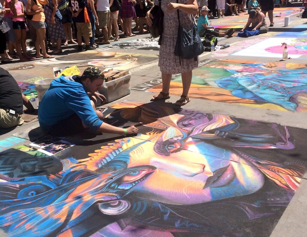 Artist working on huge, colorful sidewalk chalk art picture of woman surrounded by beautiful colors in Larimar Square as crowd of people watches on. Larimar Square, Downtown Denver, Colorado, Chalk Art Festival during summer.