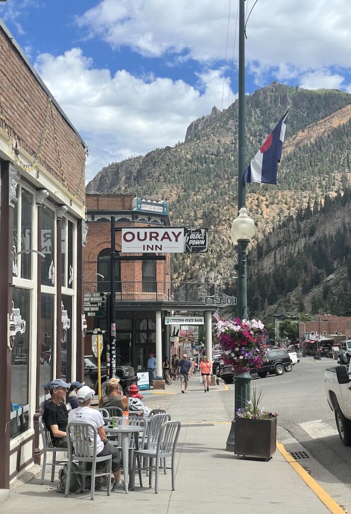 Historic brick buildings, outdoor dining table, pedestrians shopping, all surrounded by mountain views on sunny summer day. Shopping/ dining in downtown Ouray, Colorado.