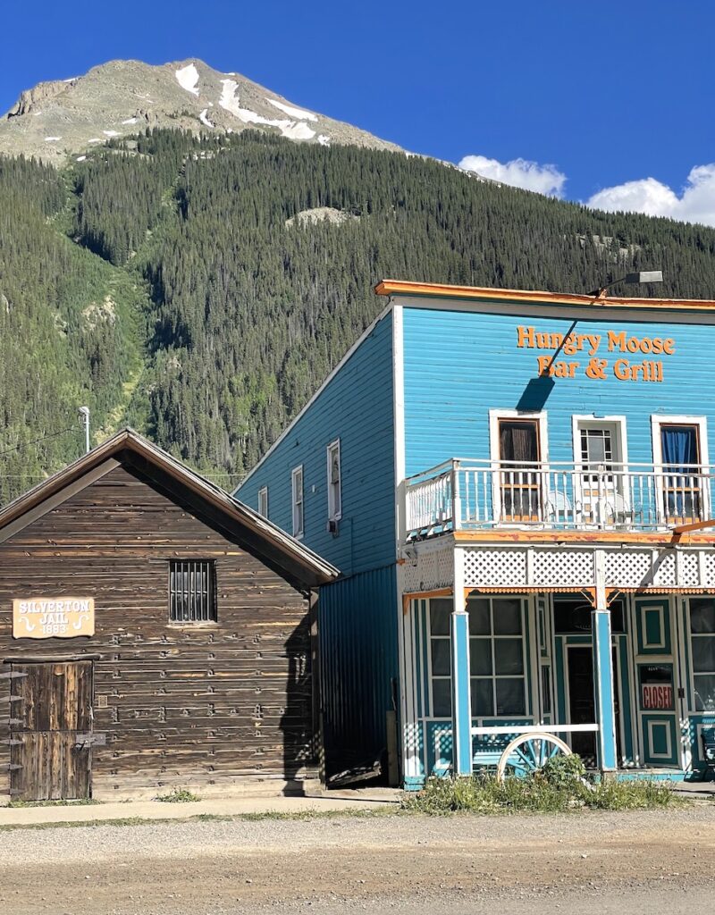 Historic mining buildings, one bright blue and one brown with dramatic mountain peak in background on sunny summer day. Silverton Jail, Hungry Moose Bar & Grill, Silverton Historic District, Silverton, Colorado.