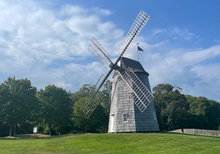 Large wooden, shingled windmill on grassy lawn with trees in background on sunny summer day. Hook Windmill, East Hampton, New York, The Hamptons.