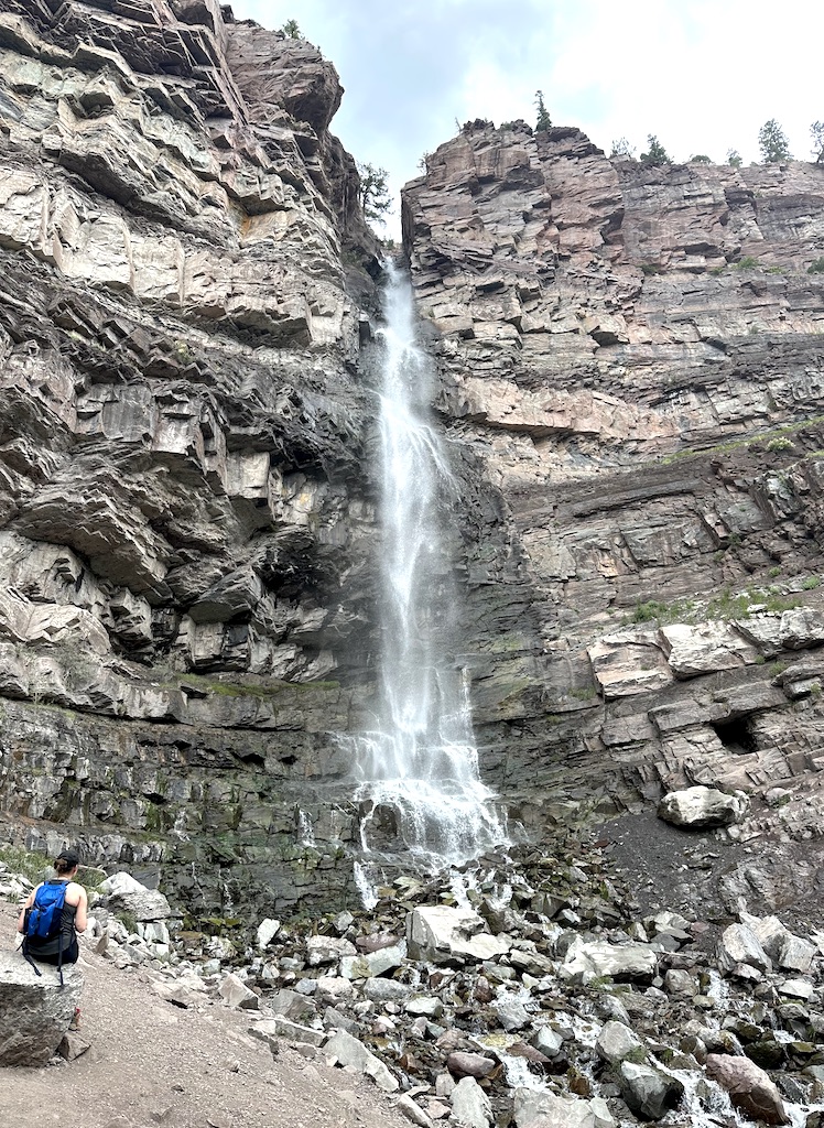 Woman sitting at base of tall free-falling waterfall surrounded by rocky cliff in summer. Lower Cascade Falls, hiking in Ouray, Colorado.