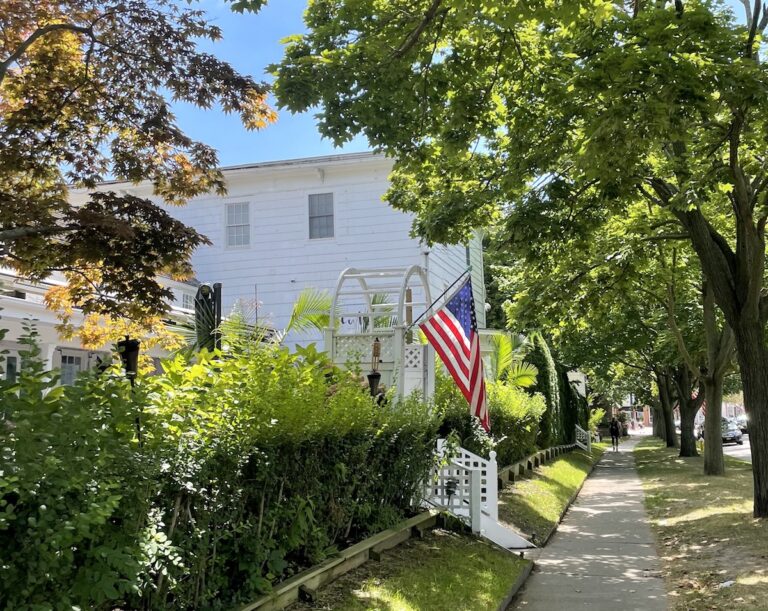 Walking through a neighborhood with white shingled homes, mature trees, and American flags on sunny summer day. Village of Southampton, Southhampton, New York, The Hamptons.