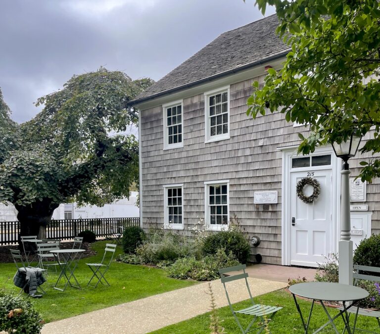 Grey shingled house with white door and windows, simple bistro table out on front lawn. Main Street, Amagansett, New York, The Hamptons.