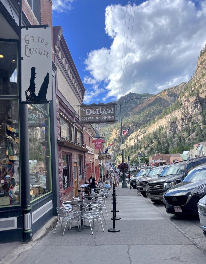 Historic brick buildings, outdoor dining tables, all surrounded by mountain views on sunny summer day. Shopping/ dining in downtown Ouray, Colorado.