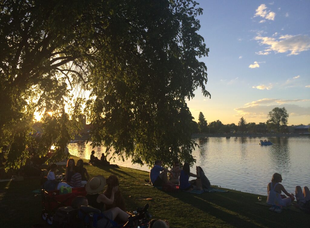Crowd sitting in grassy lawn surrounded by tall trees overlooking calm pond during sunset music event. City Park Jazz, Jazz at the Park, City Park, Denver, Colorado.
