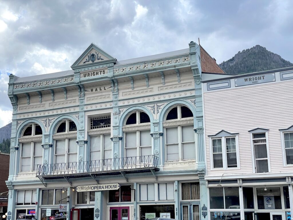 Ornate iron building facade in teal and white built in 1880 with mining-era building next door and mountain views in background. Wright Opera House, Downtown Ouray, Ouray, Colorado.