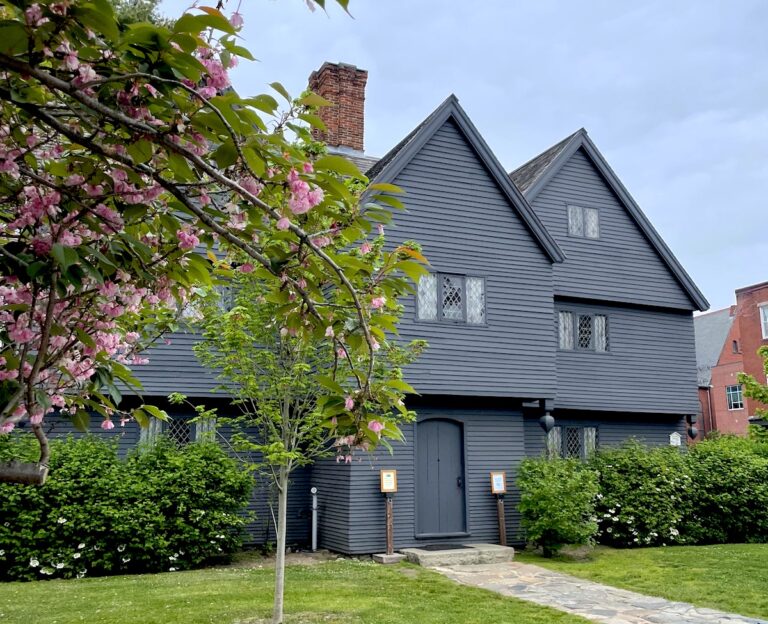 Black, shingled gothic home on small lot with green gras and simple shrubs, cherry blossom tree in foreground on gloomy day. Salem Witch House, Salem, Massachusetts.