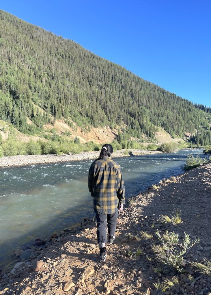 Woman standing in front of river confluence with mountains in background on sunny summer evening. Confluence of Cement Creek and Animas River, Silverton, Colorado.