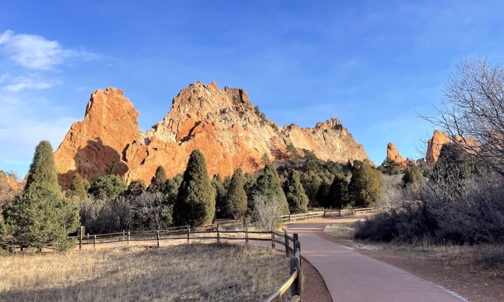 Huge red rocks jutting up from earth with paved path winding towards unique landscape surrounded by trees on sunny day. Garden of the Gods, Colorado Springs, Colorado.