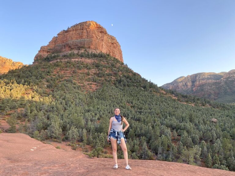 Woman smiling for camera in front of red rock mountain with lower portion of mountain covered in green evergreen pines on sunny evening. Pink Jeep Tours, Broken Arrow Route at sunset, Sedona, Arizona.