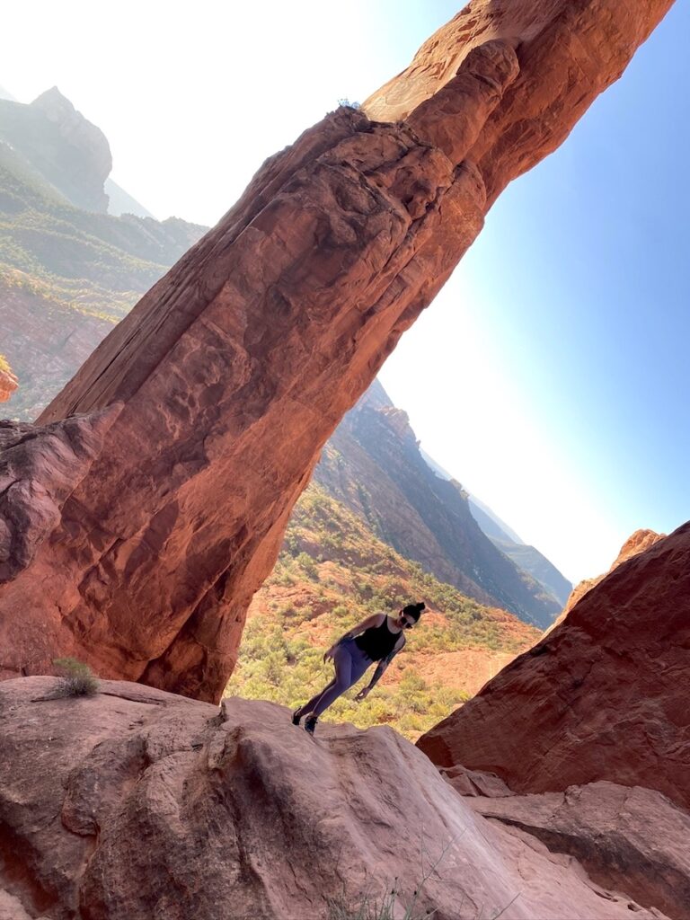 Woman hiking between two huge red rock spires with dramatic desert landscape in background on sunny morning. Cathedral Rock Trail, Sedona, Arizona.