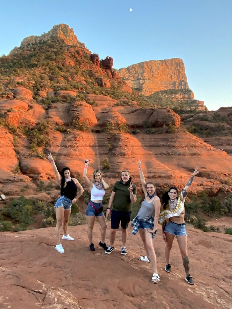 Group of friends smiling and posing for camera in front of red rock mountains during sunset. Pink Jeep Tours, Broken Arrow Route at sunset, Sedona, Arizona.