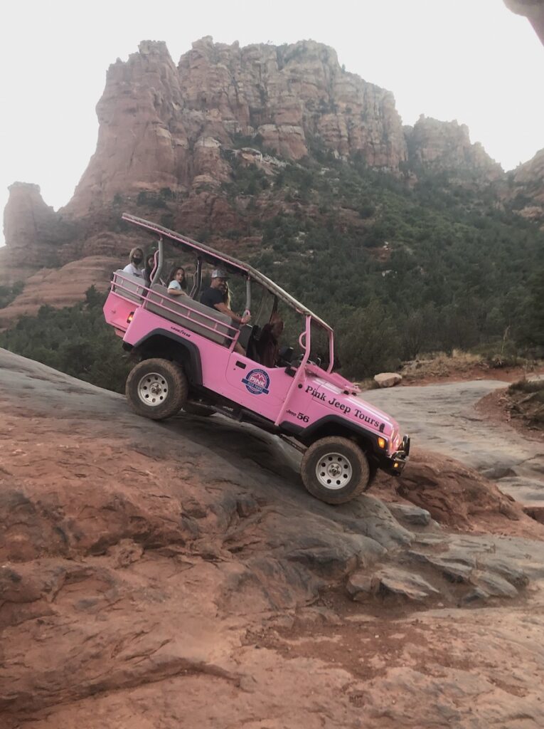 Pink jeep riding steep slick red rock with red rock mountains in background at sunset. Pink Jeep Tours, Broken Arrow Route at sunset, Sedona, Arizona.