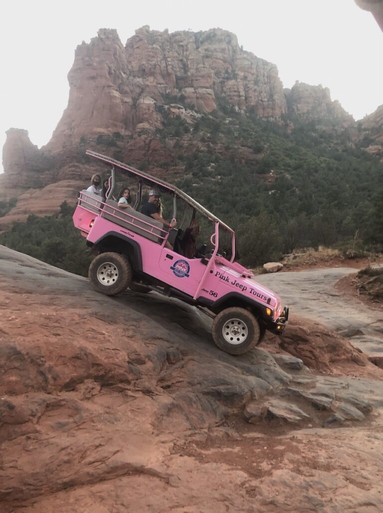 Pink jeep riding steep slick red rock with red rock mountains in background at sunset. Pink Jeep Tours, Broken Arrow Route at sunset, Sedona, Arizona.