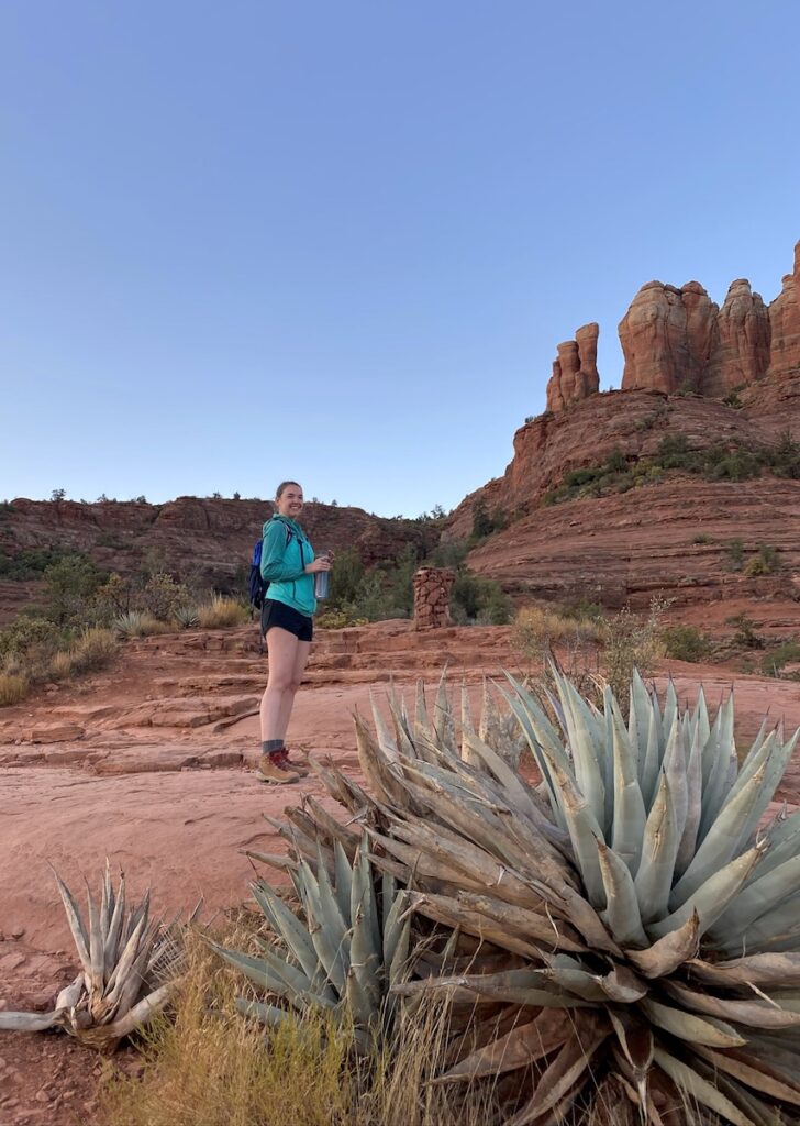 Woman smiling at camera with large agave plant in foreground and red rock spires in background during early morning. Cathedral Rock Trail, Sedona, Arizona.