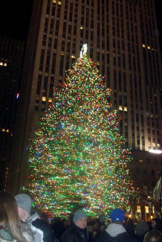 Crowd of people around huge outdoor Christmas tree covered in colorful lights with skyscraper buildings in background. The Rockefeller Center Christmas Tree, New York City, Manhattan, NYC, New York.