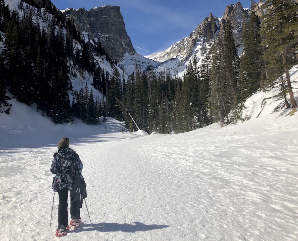 Women snowshoe hiking on frozen lake surrounded by incredible towering mountain views on sunny winter day. Dream Lake hike, winter in Rocky Mountain National Park, Estes Park, Colorado. Things to do in Estes Park.