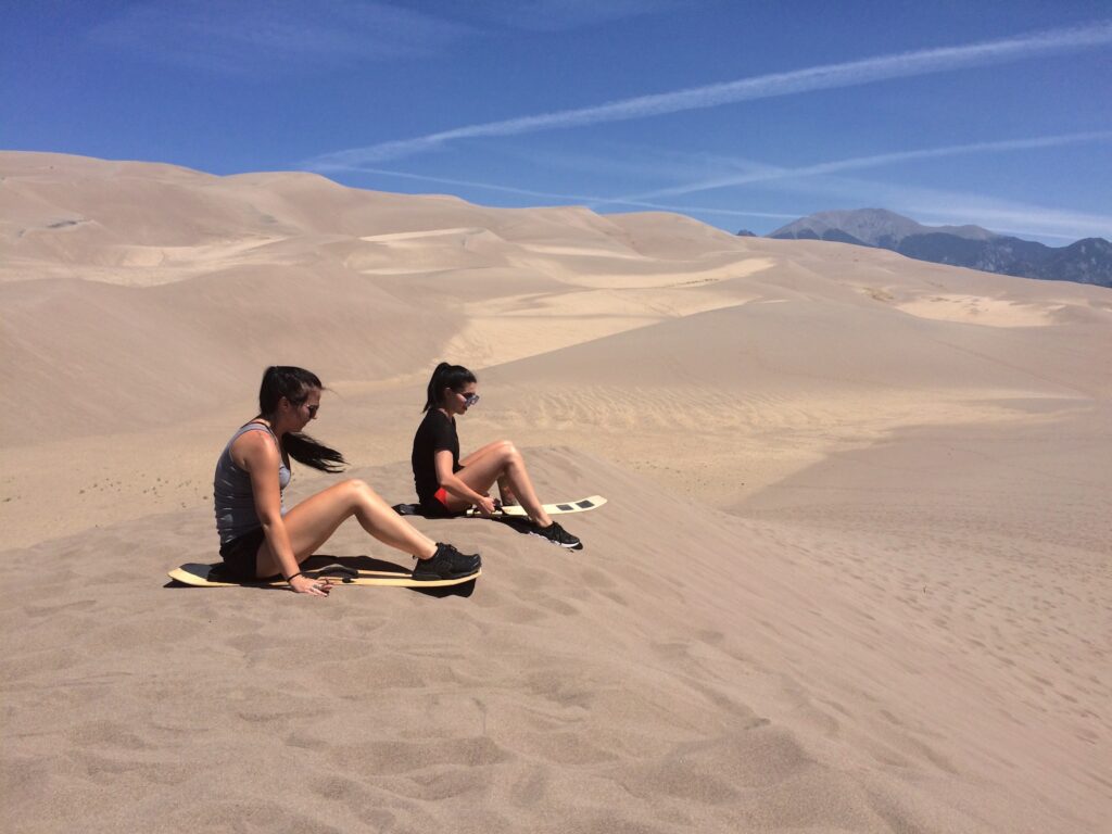Two women sitting on sand sleds ready to sled down sand dune, in background are huge sand dunes and distance mountains on sunny summer day. Great Sand Dunes National Park, Alamosa, Colorado.