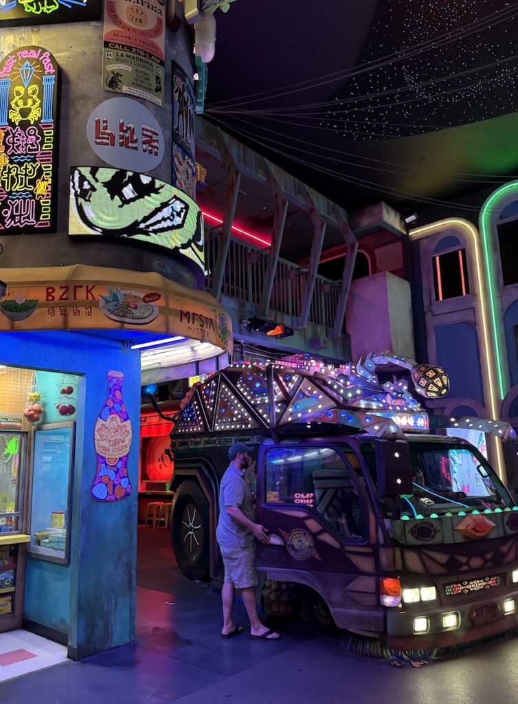 Man getting into neon van surrounded by replica city in other-worldly interactive art exhibit. Meow Wolf Denver Convergence Station, Denver, Colorado.