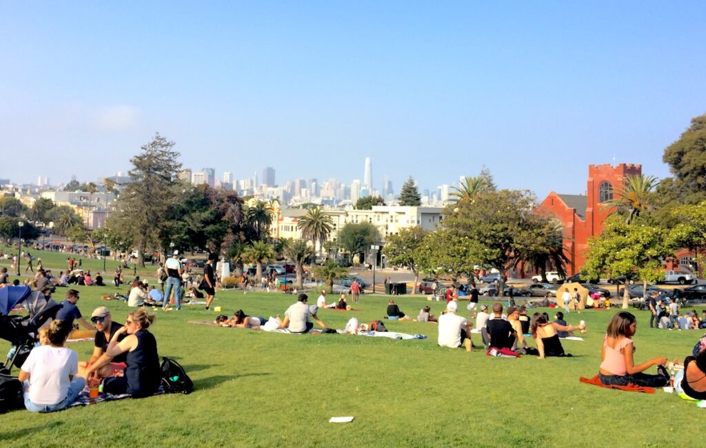 Large grassy lawn full of people lounging out, picnicking, and playing games, lawn overlooks San Francisco skyline. Mission Dolores Park, San Francisco, California.