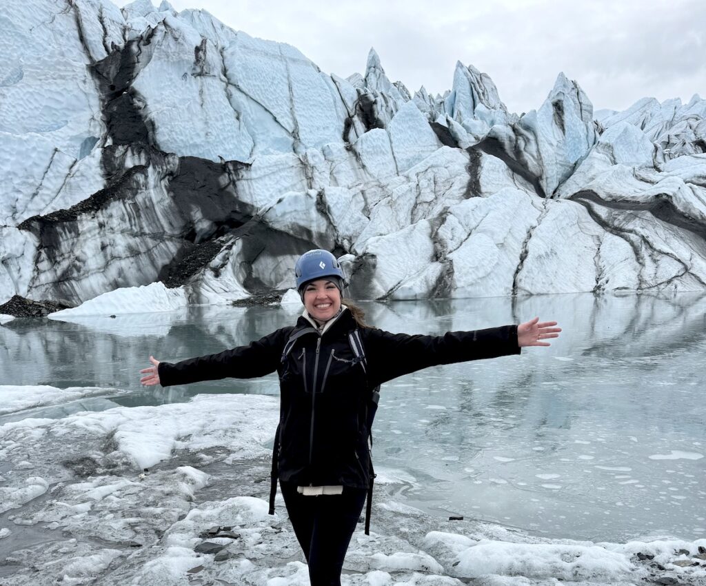 Woman smiling while standing on glacier with jagged stripped glacier ice and small glacier lake in background on gloomy day. Matanuska Glacier, near Anchorage, Alaska. (horizontal)