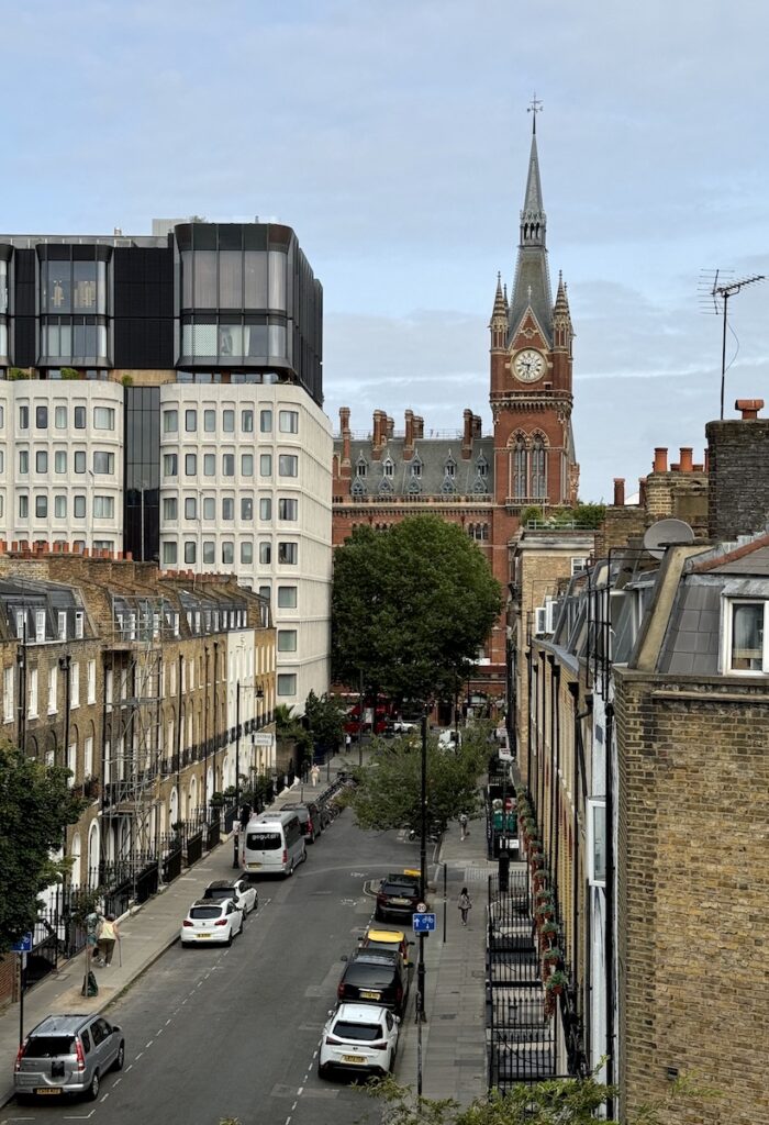 Cityscape with large red brick clock tower and spire rising up behind buildings on city street. Kings Cross Station, London, England.